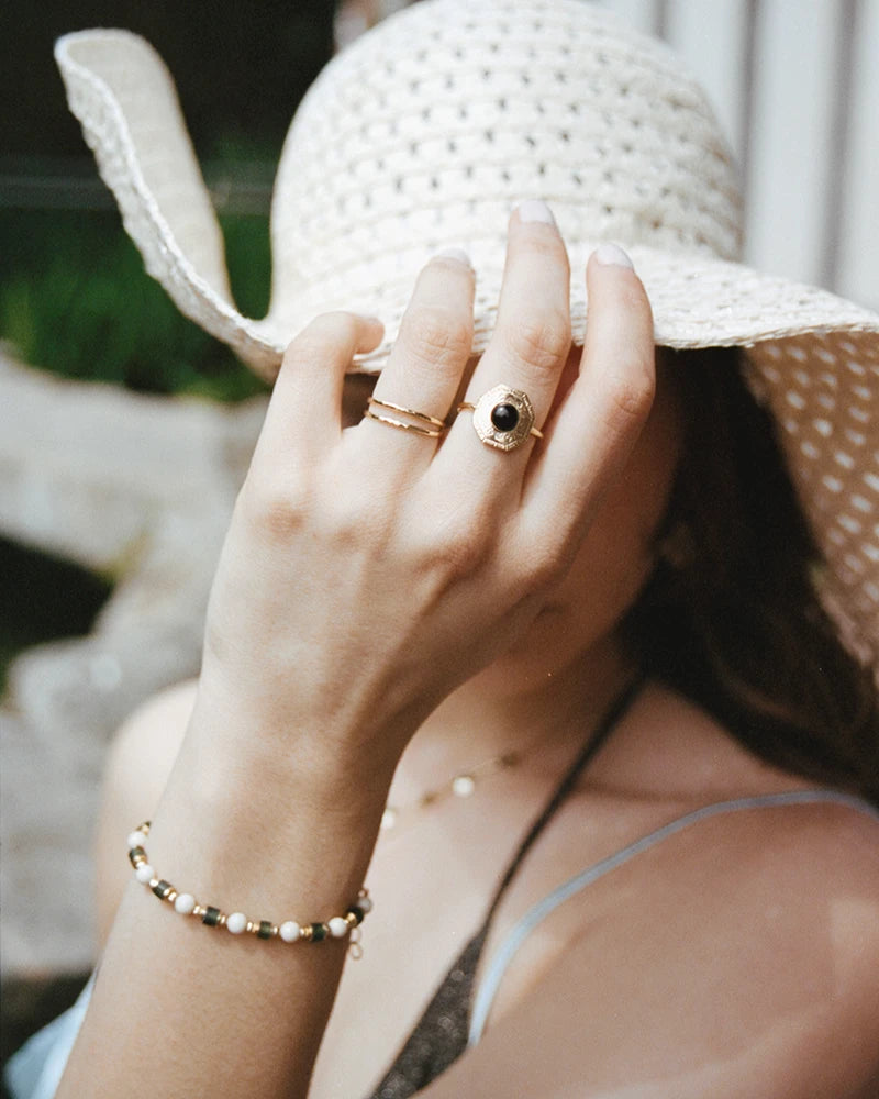Zoom sur le bracelet Sanjula porté avec élégance, en harmonie avec une bague dorée et un chapeau de paille d’été.