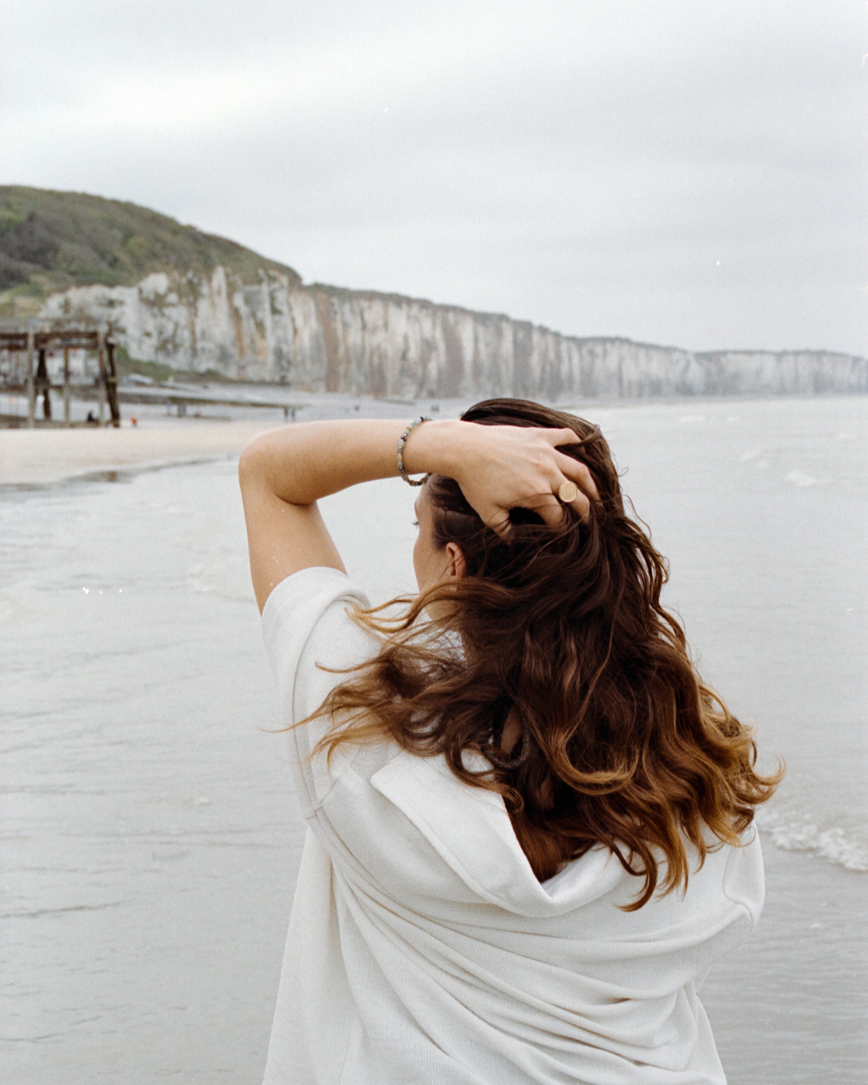 Vue arrière d'une femme au bord de la mer, cheveux au vent, portant le bracelet Urja devant les falaises normandes, esprit poétique et naturel.