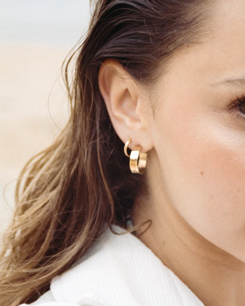 Vue rapprochée d’une oreille ornée de la créole Sundari dorée, finition martelée à la main, avec lumière naturelle de plage.