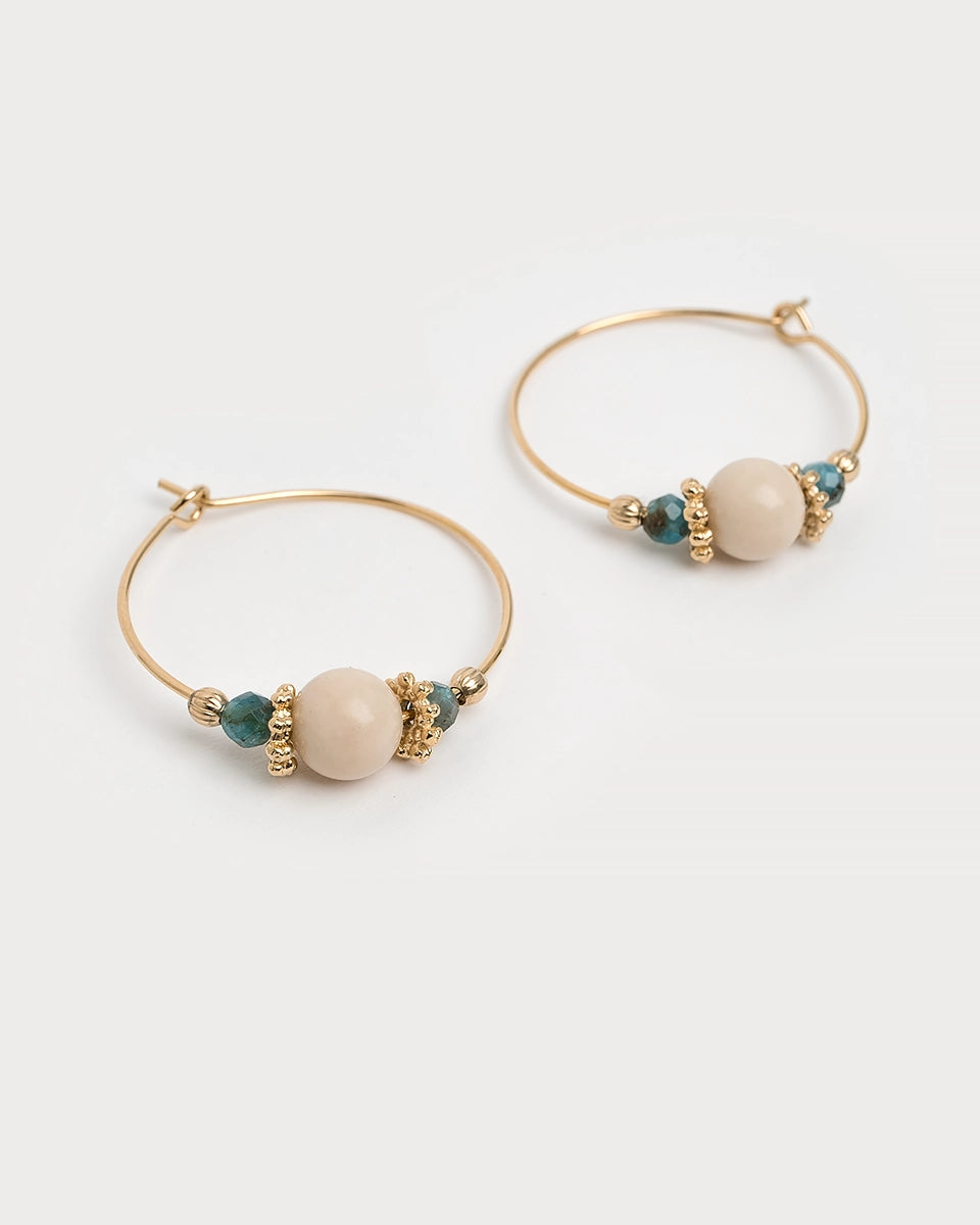 Paire de boucles d’oreilles créoles Svara dorées, ornées de perles d’apatite bleue et de pierre fossile beige, posées à plat sur fond blanc.