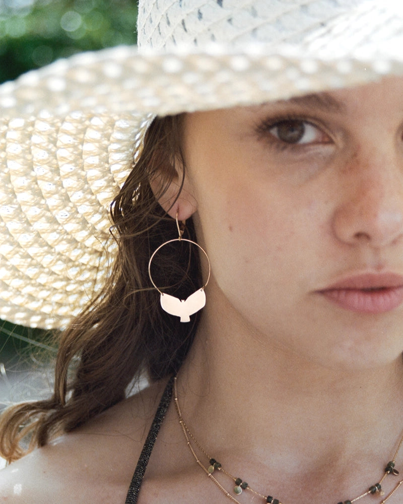 Femme portant les boucles d’oreilles oiseau dorées Udaan, créoles fines avec forme d’oiseau découpée à la main, photographiée à la lumière naturelle sous un chapeau de paille.