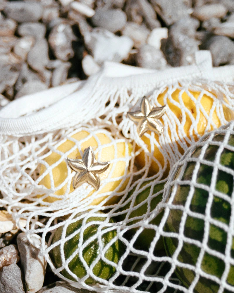 Paire de boucles d’oreilles dorées en forme d’étoile de mer JAL, posées sur un filet en coton avec citrons et fruits en arrière-plan.