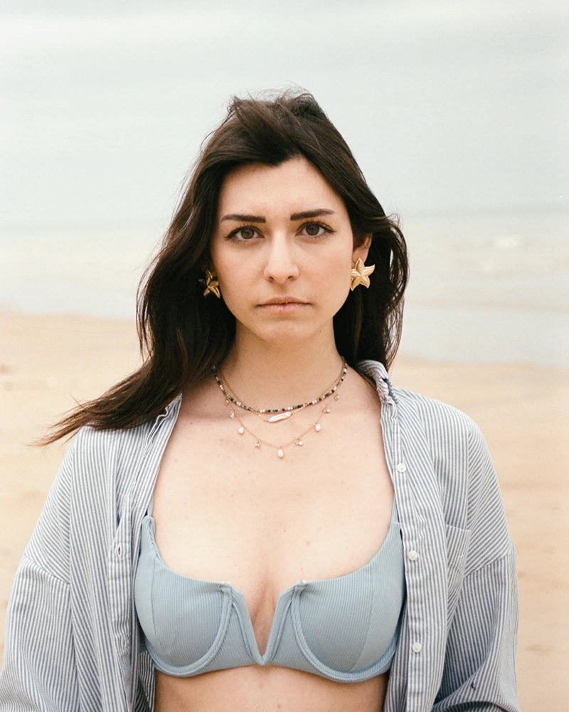 Femme sur la plage portant les boucles d’oreilles dorées JAL en forme d’étoile de mer, avec collier de perles et chemise rayée ouverte.