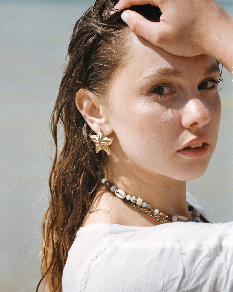 Portrait d’une femme aux cheveux mouillés, portant les boucles d’oreilles dorées Jal étoile de mer, avec un collier à coquillages.