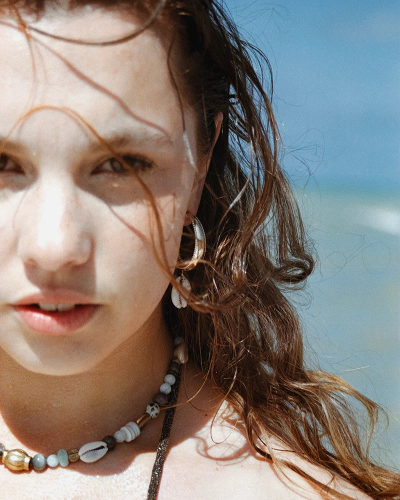 Portrait d’une femme en bord de mer portant la boucle d’oreilles coquillage Samudra, créole dorée avec cauri naturel, associée à un collier de perles colorées.