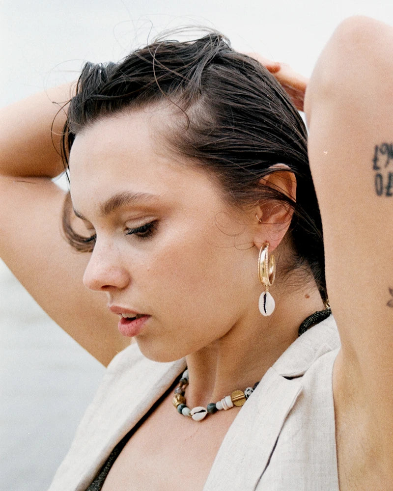 Femme vue de profil portant la boucle d’oreilles coquillage Samudra, créole dorée avec pendentif cauri, accompagnée d’un collier en perles naturelles aux tons marins.