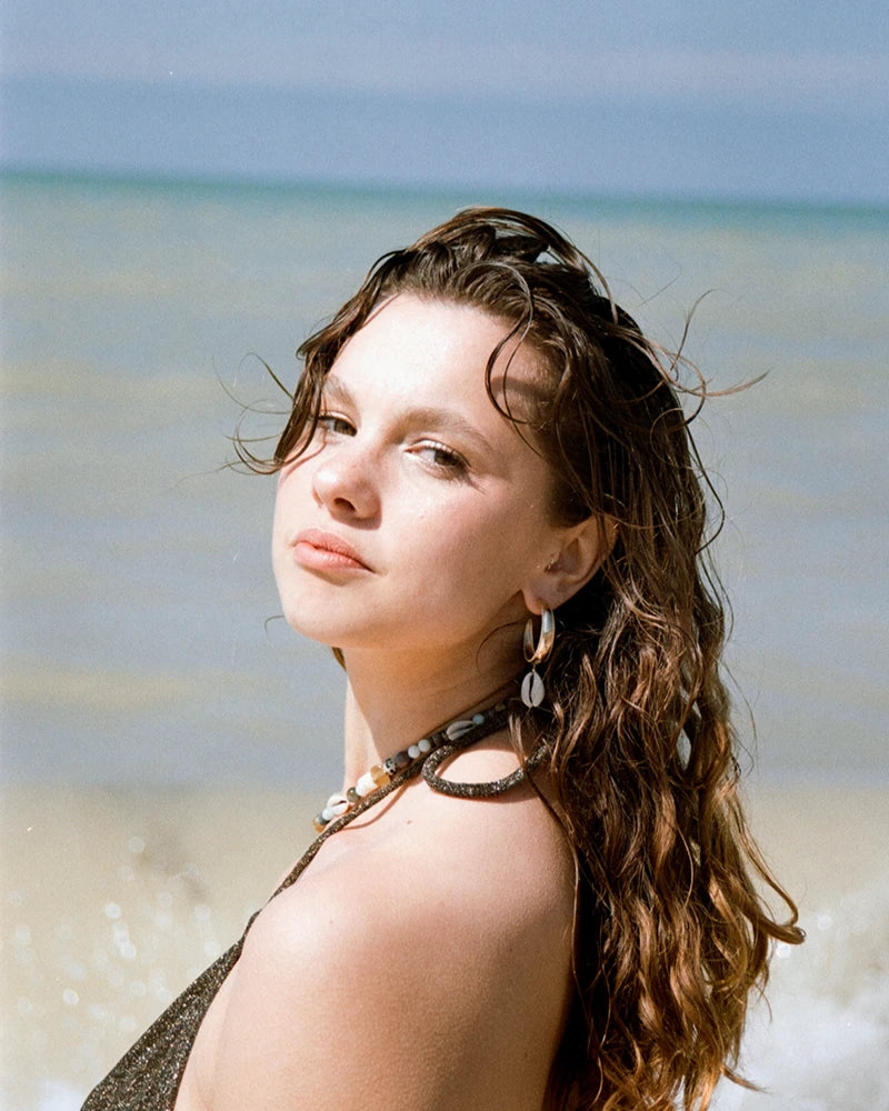 Femme portant la boucle d’oreilles coquillage Samudra de Gauhart, créole dorée et cauri naturel, photographiée en bord de mer dans une lumière estivale.