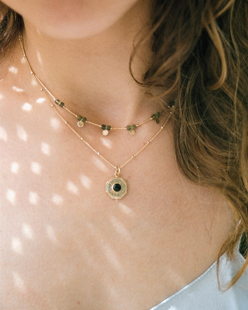 Collier Gatika porté en accumulation, mettant en valeur le médaillon doré à l’onyx noir sur une peau délicatement éclairée par le soleil.