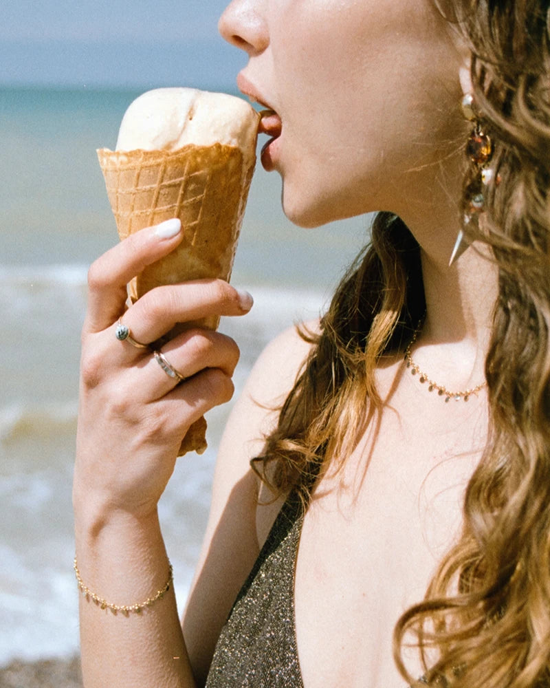 Femme dégustant une glace sur la plage, portant le collier doré Nimisha en laiton plaqué or, bijou délicat et lumineux.