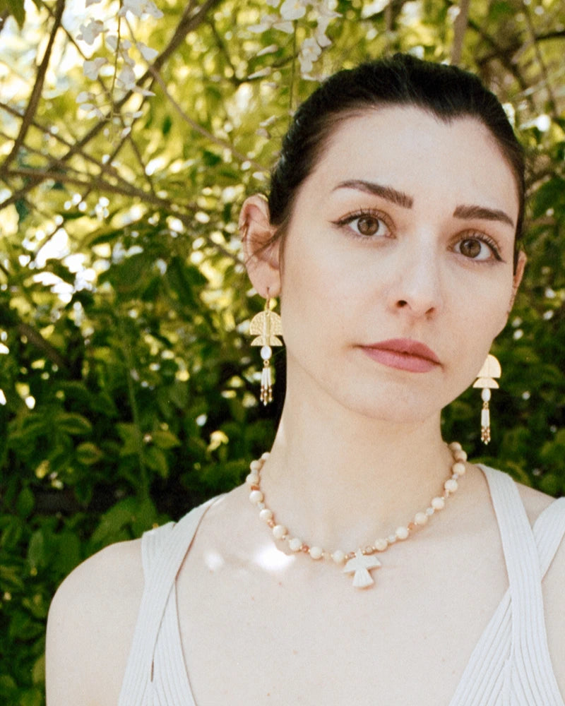 Femme portant les boucles d’oreilles dorées à pampilles Gauhart et un collier perlé avec pendentif oiseau, photographiée devant un feuillage lumineux.