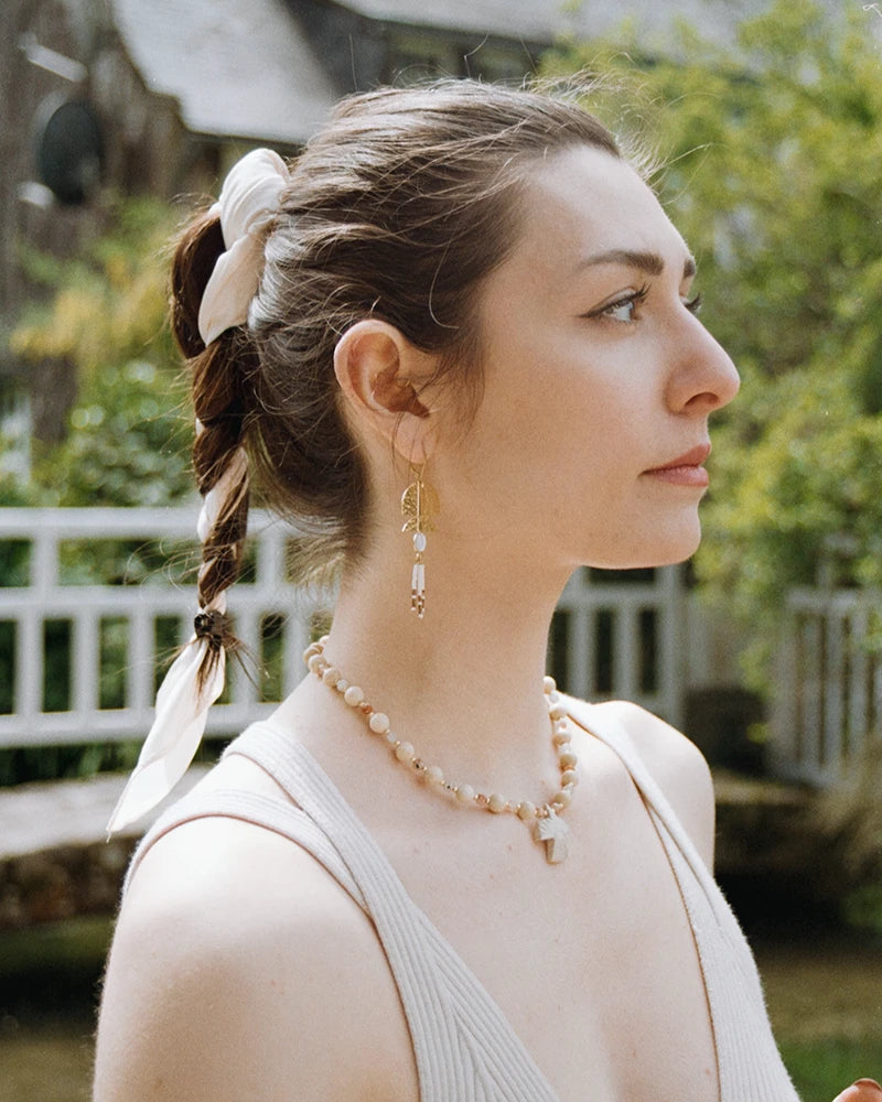 Femme de profil portant le collier Minali en pierre de lune et pierre fossile, assorti à une boucle d’oreille dorée, dans un jardin verdoyant.