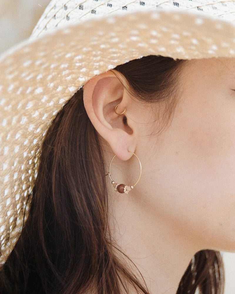 Femme portant une boucle d’oreilles créole dorée Svara avec perles de pierre de lune et d’agate, photographiée à la lumière naturelle sous un chapeau de paille.