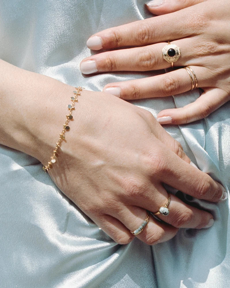 Poignet féminin sur fond de tissu satiné bleu ciel, mettant en valeur le bracelet doré à pampilles et des bagues dorées raffinées.