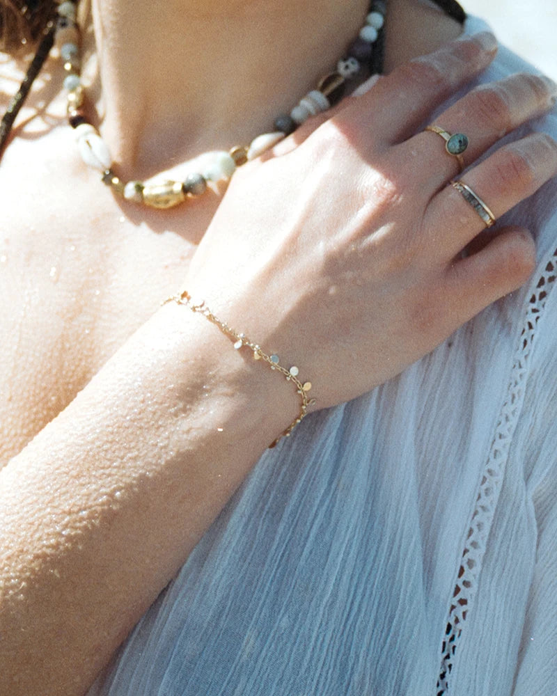 Poignet orné du bracelet doré à pampilles capturé en extérieur, lumière naturelle sur peau mouillée et robe fluide estivale, ambiance bord de mer.