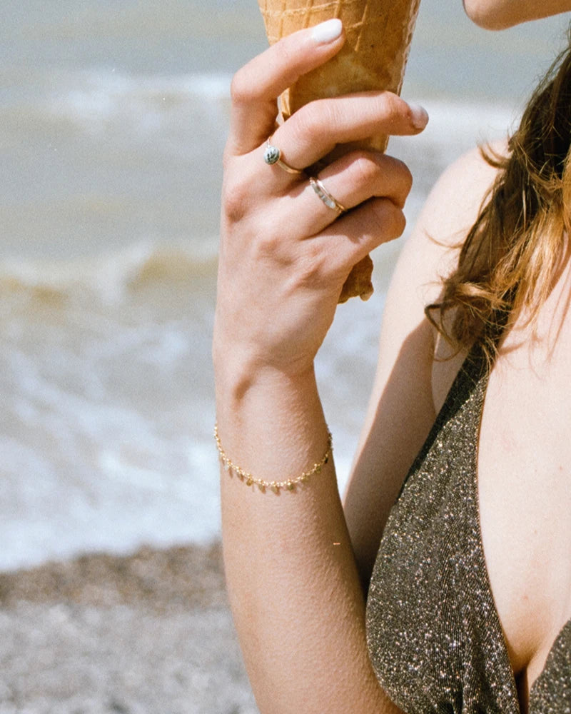 Bracelet doré à pampilles porté à la plage, cône glacé à la main, look estival naturel et lumineux.
