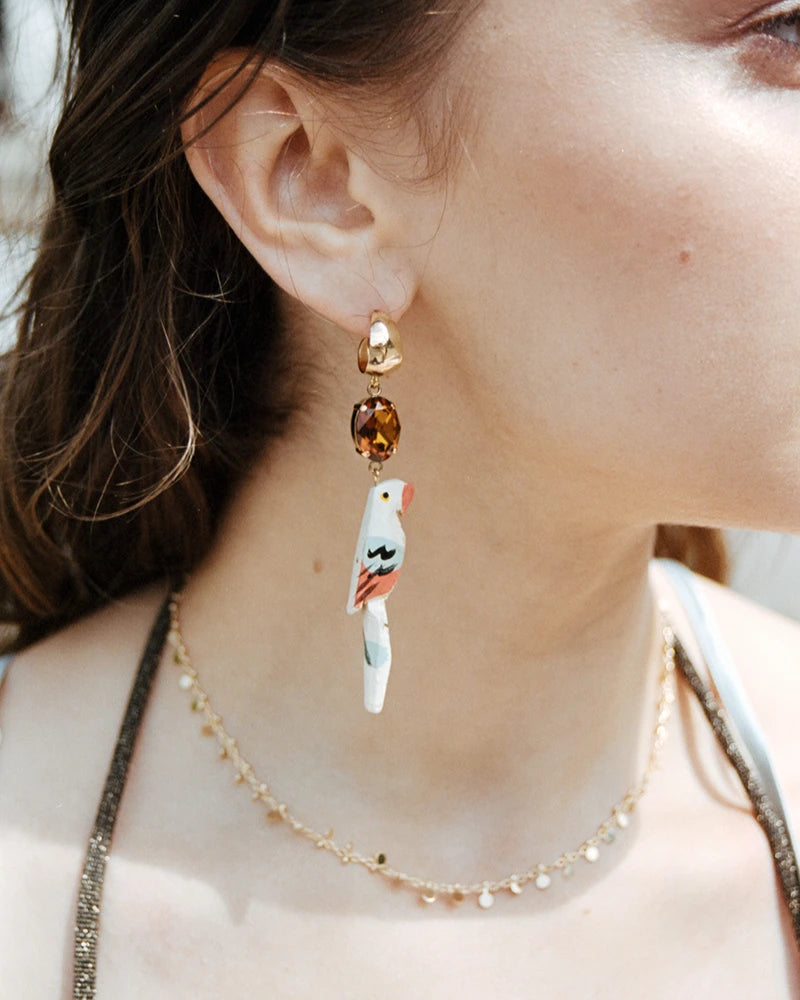 Femme portant une boucle d’oreille dorée avec pendentif perroquet coloré suspendu à une pierre ovale ambrée, mise en valeur par un collier fin doré.