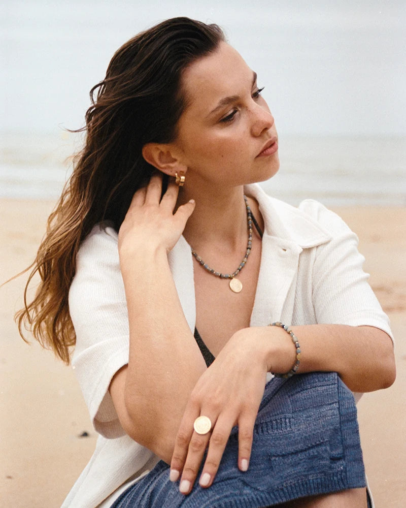 Femme assise sur une plage, portant le bracelet Urja associé à d'autres bijoux dorés, dans une ambiance estivale élégante au bord de l’eau.