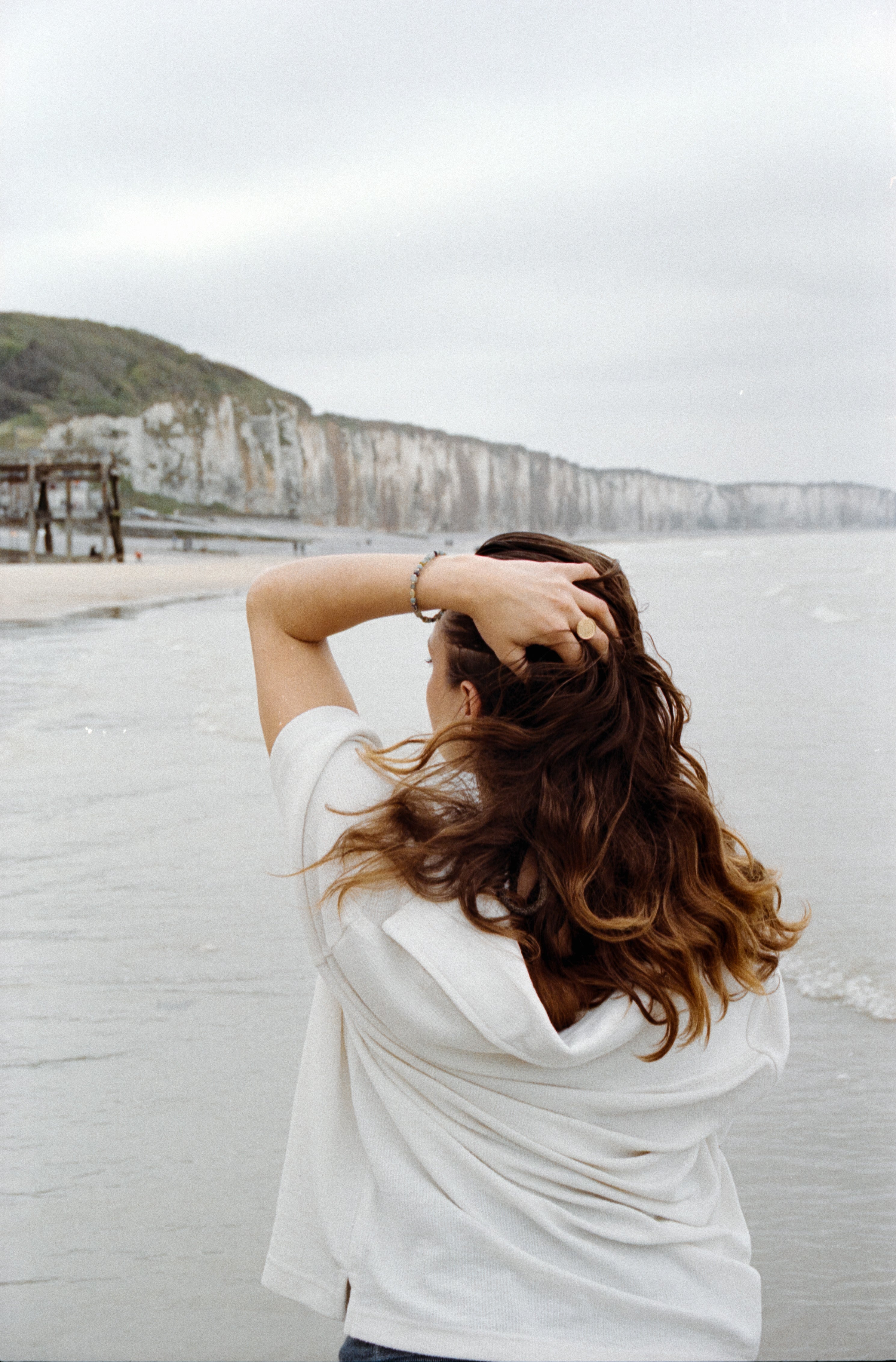 Vue arrière d'une femme au bord de la mer, cheveux au vent, portant le bracelet Urja devant les falaises normandes, esprit poétique et naturel.